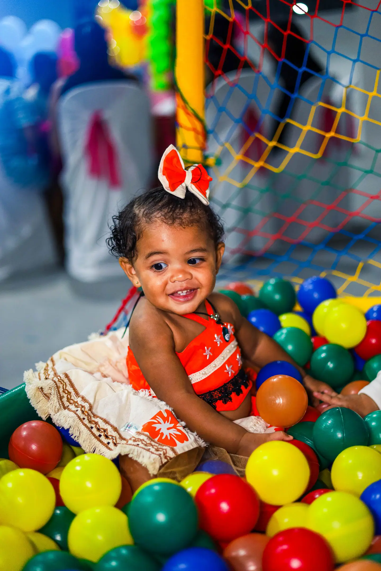 Bebê com laço vermelho sorri numa piscina de bolinhas coloridas