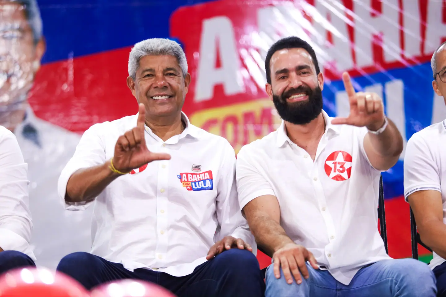 Dois homens de camisa branca riem e gesticulam sentados no palco