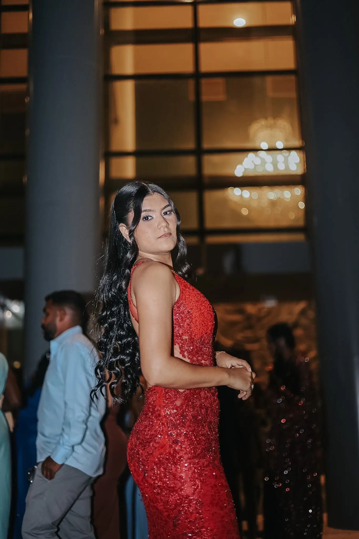 Jovem de vestido vermelho bordado posa entre colunas e lustres